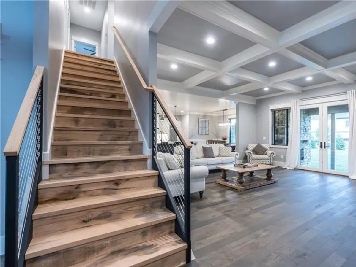 Wooden staircase with black metal railing leads to a living room with gray walls and a coffered ceiling.
