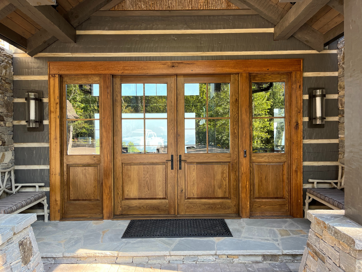 Wooden double doors with glass panels and sidelights on a stone porch, flanked by sconces and seating.