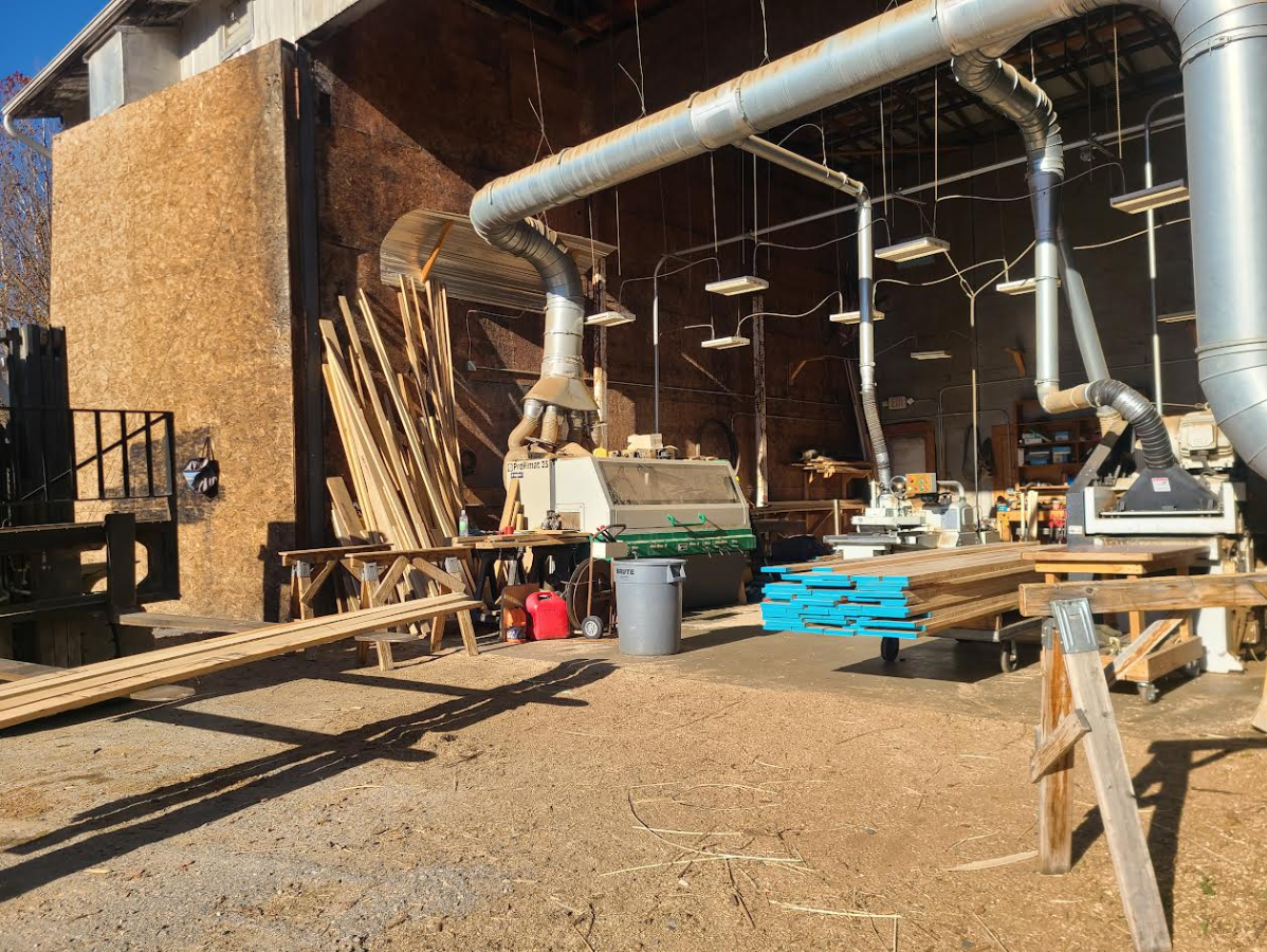 Exterior view of a woodworking shop with lumber, ventilation pipes, and tools under a partially covered structure.