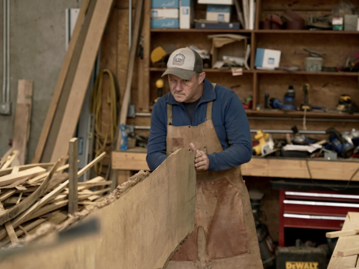 Carpenter in apron working with a large wooden plank in a workshop with tools and shelving.