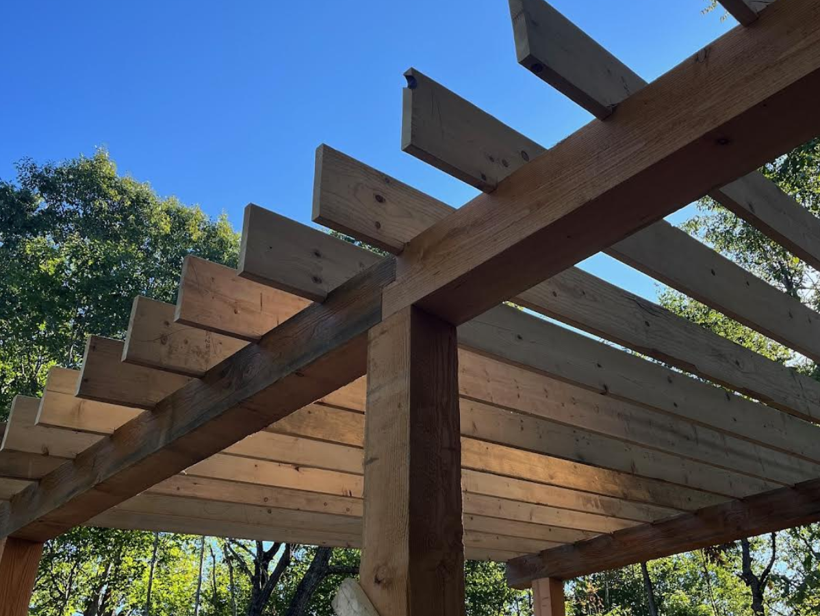 Wooden pergola under construction against a blue sky, with horizontal beams and vertical posts.