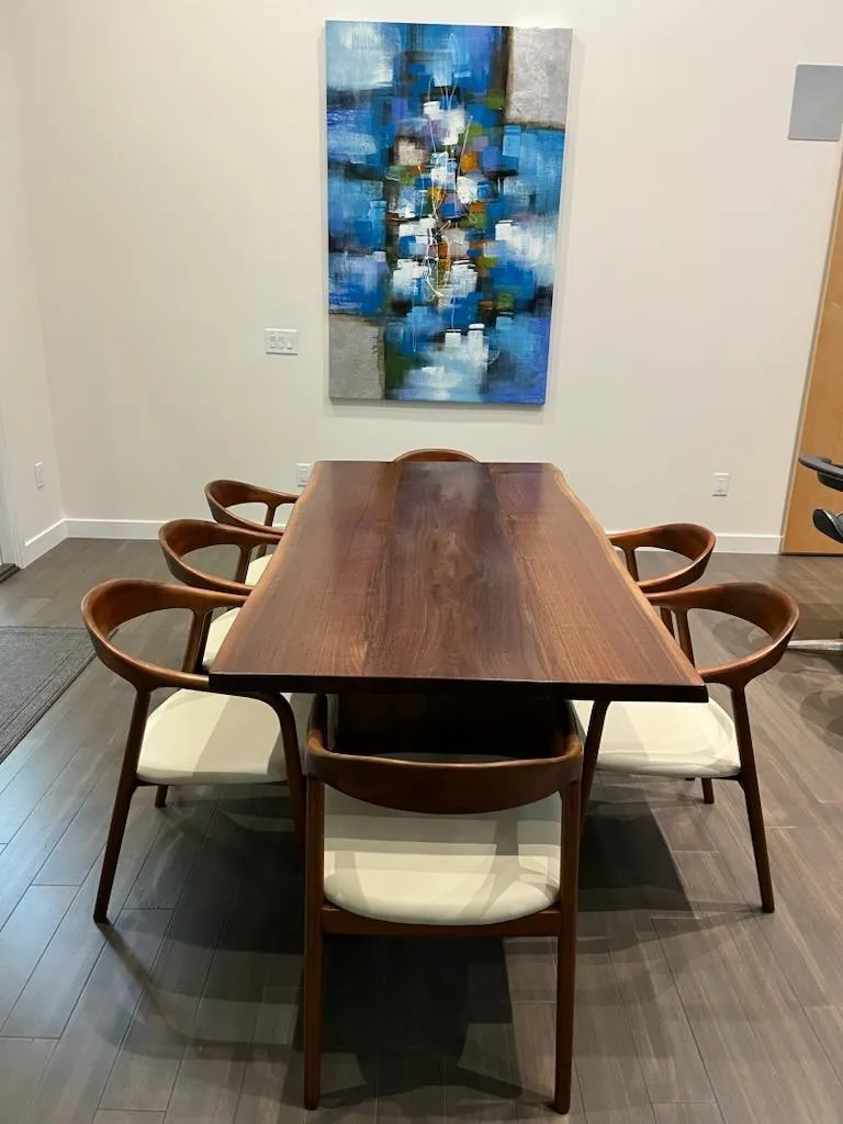 Wooden dining table with six chairs, white cushions, under abstract blue art in a modern room.