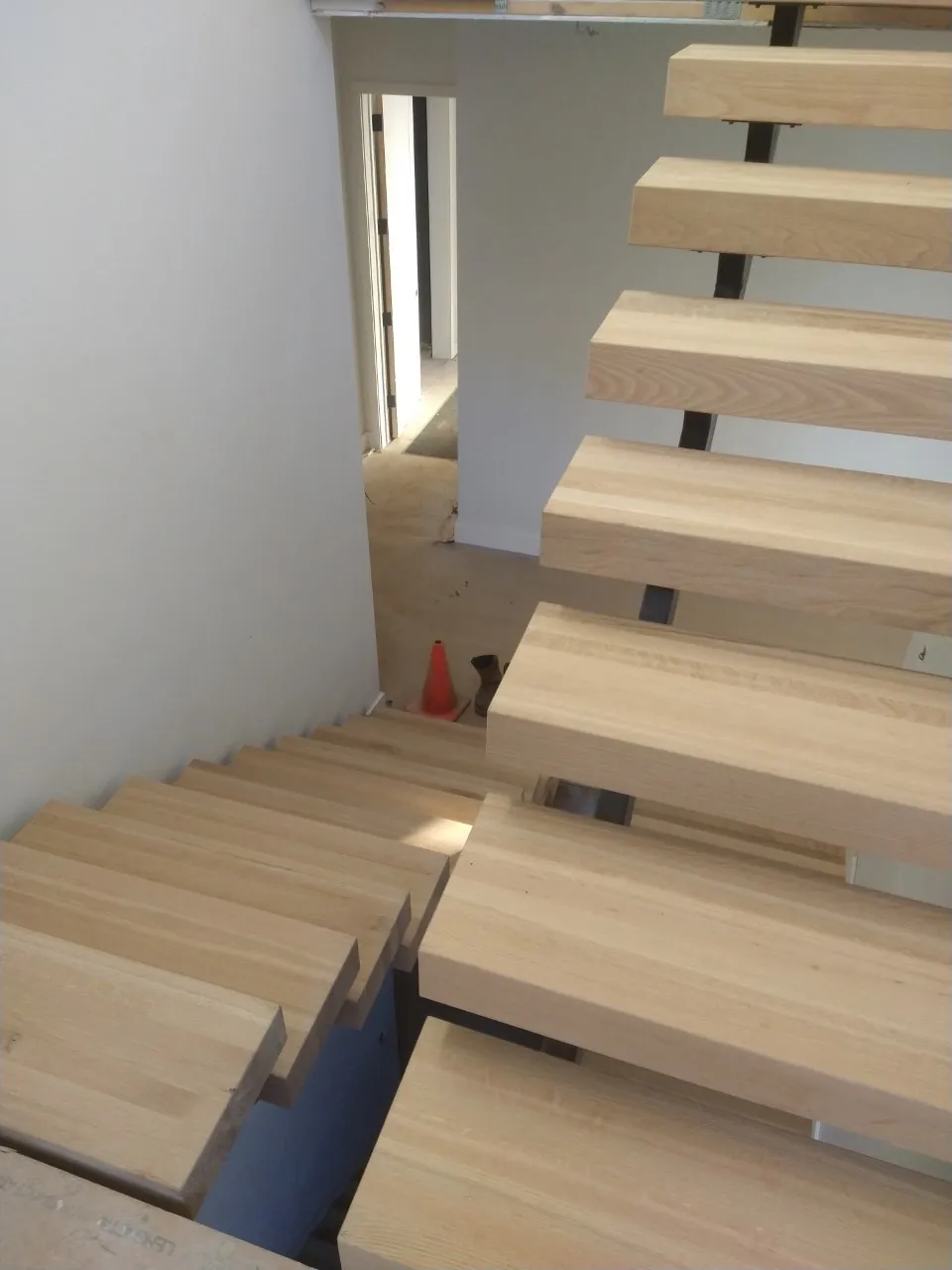 Wooden staircase with open treads and dark metal supports; natural light streams in.