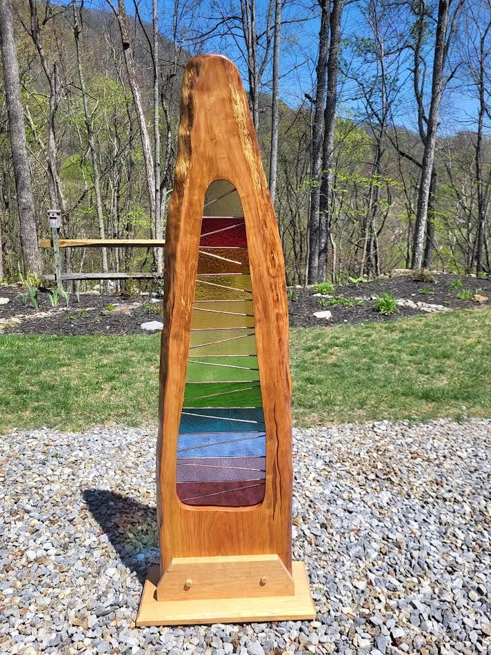 Tall wooden sculpture with colored glass panels in a natural outdoor setting.
