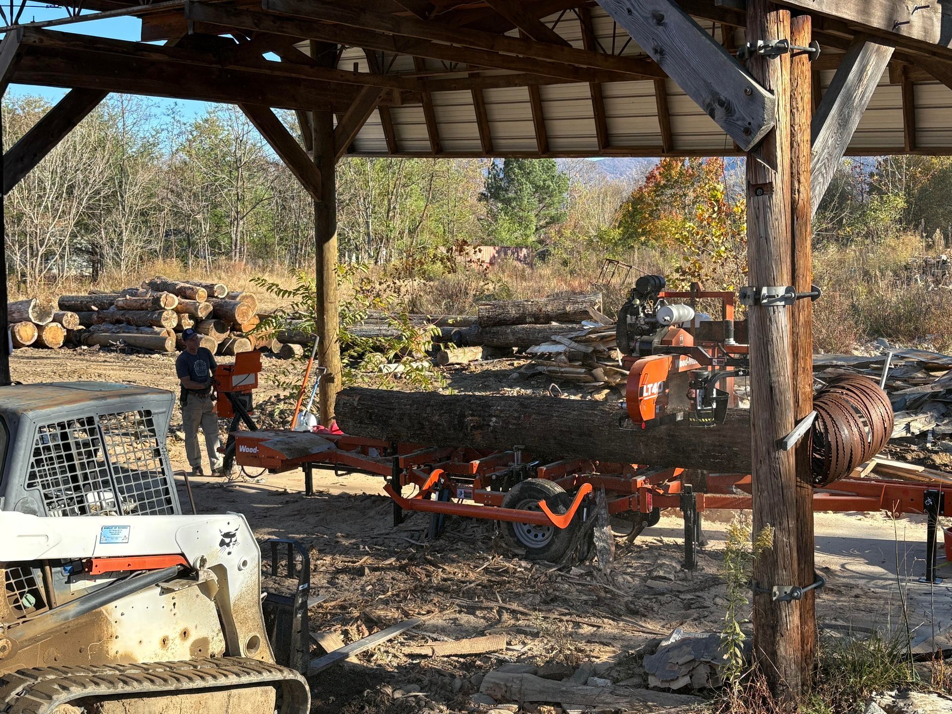 Men sawing a log on a sawmill under a wooden shelter, logs in the background, a small tractor in the foreground.