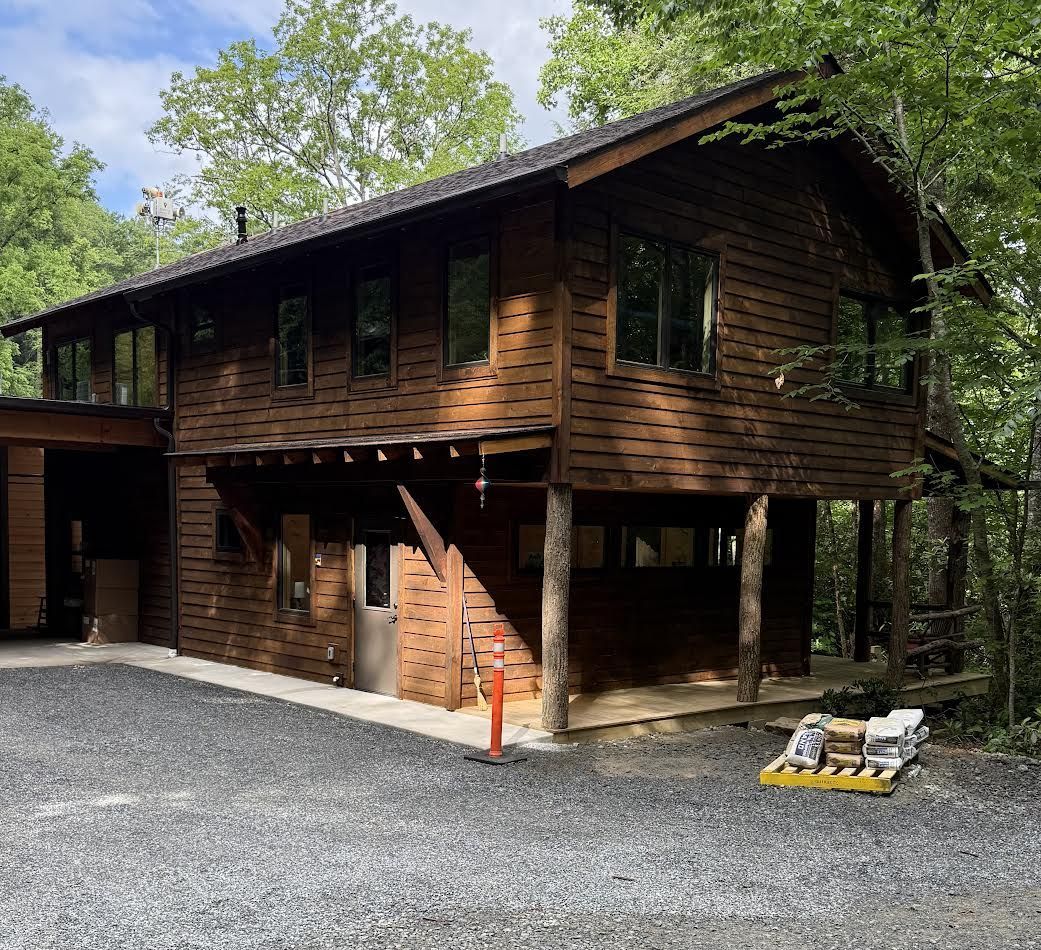 Two-story brown wooden cabin with gravel driveway, surrounded by trees.