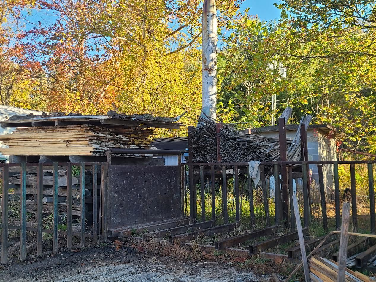 A fenced area with stacked wood, a cylindrical structure, and fall foliage in the background.