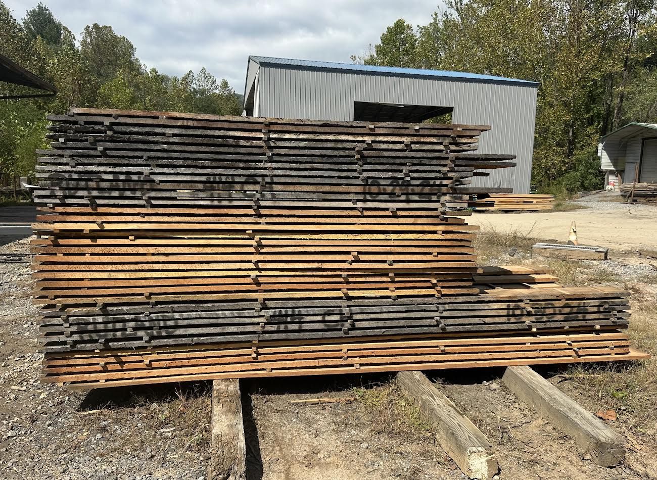 Stack of weathered wooden planks at a lumberyard, with a corrugated metal shed in the background.