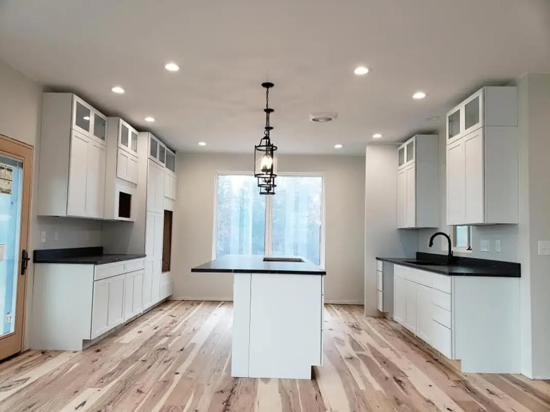Modern kitchen with white cabinets, black countertops, and wood flooring; centered island with light fixture.