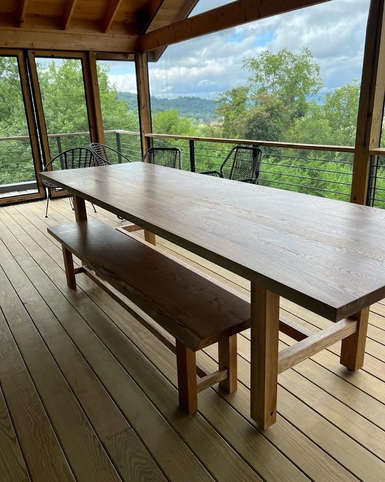 Wooden dining table and bench on a deck overlooking a view of trees and sky.