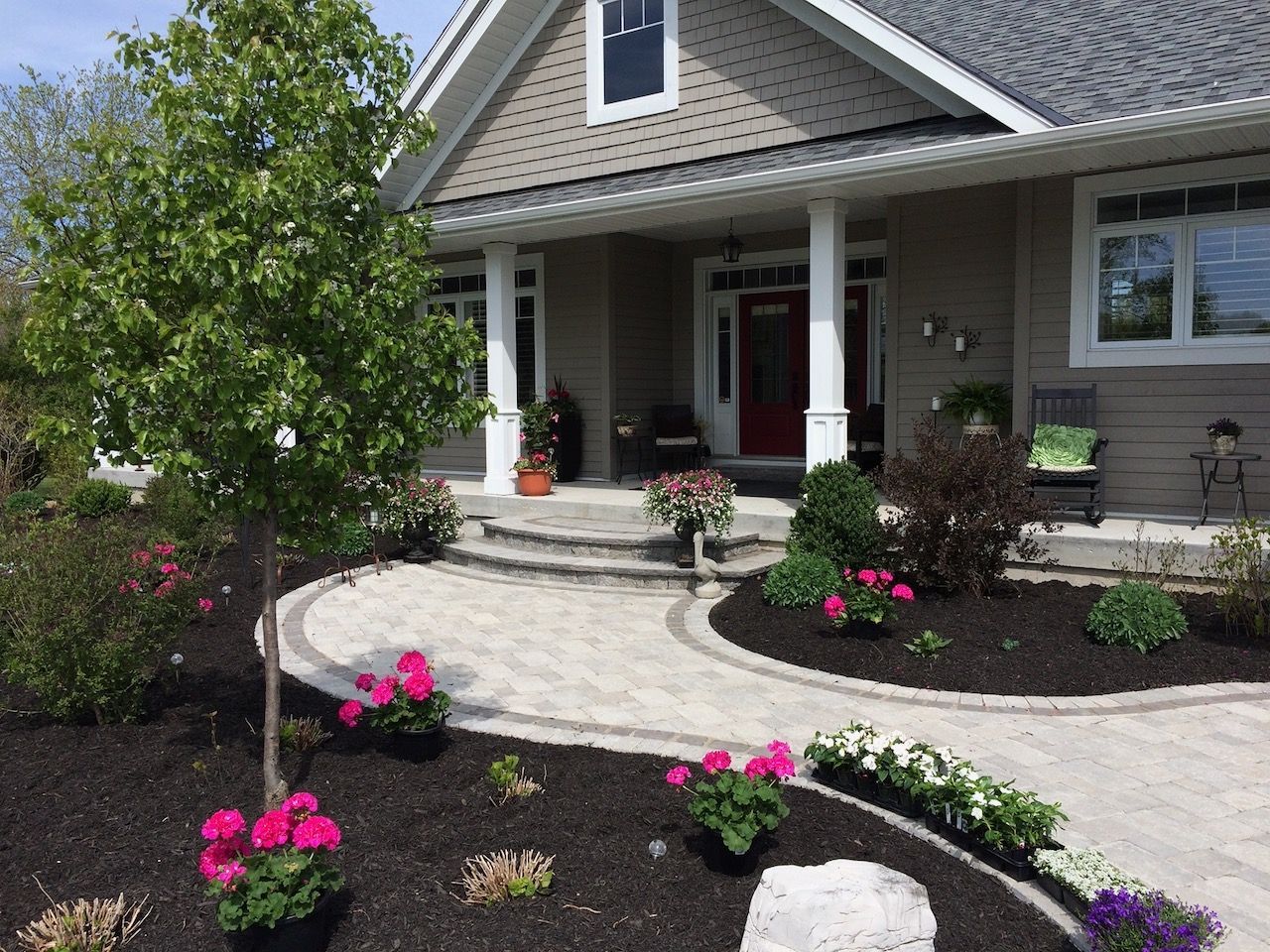 Stone pathway leads to a house with a red door, flanked by flowerbeds and a tree.
