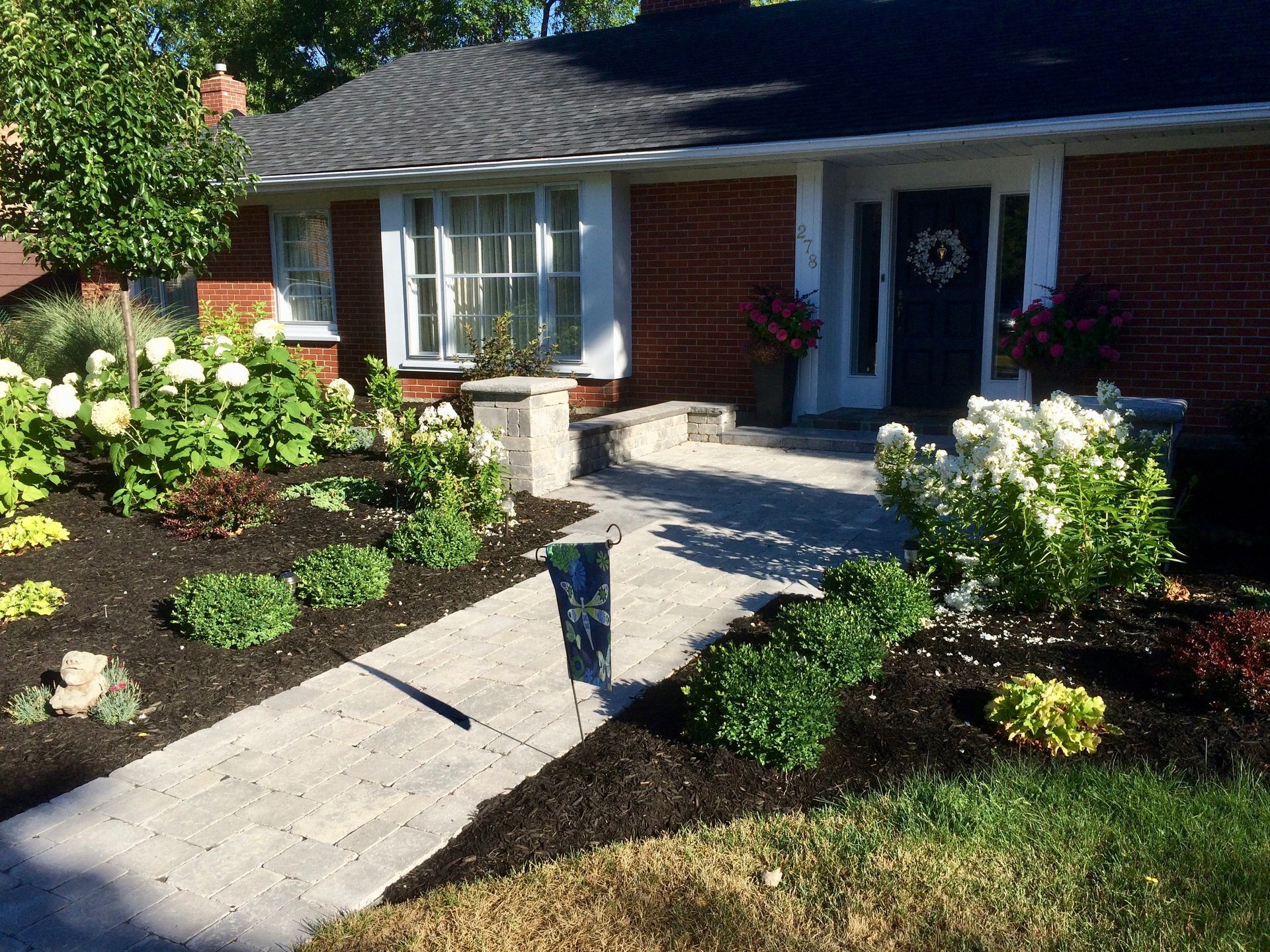 Brick house with concrete path, surrounded by flowerbeds with white and green plants.