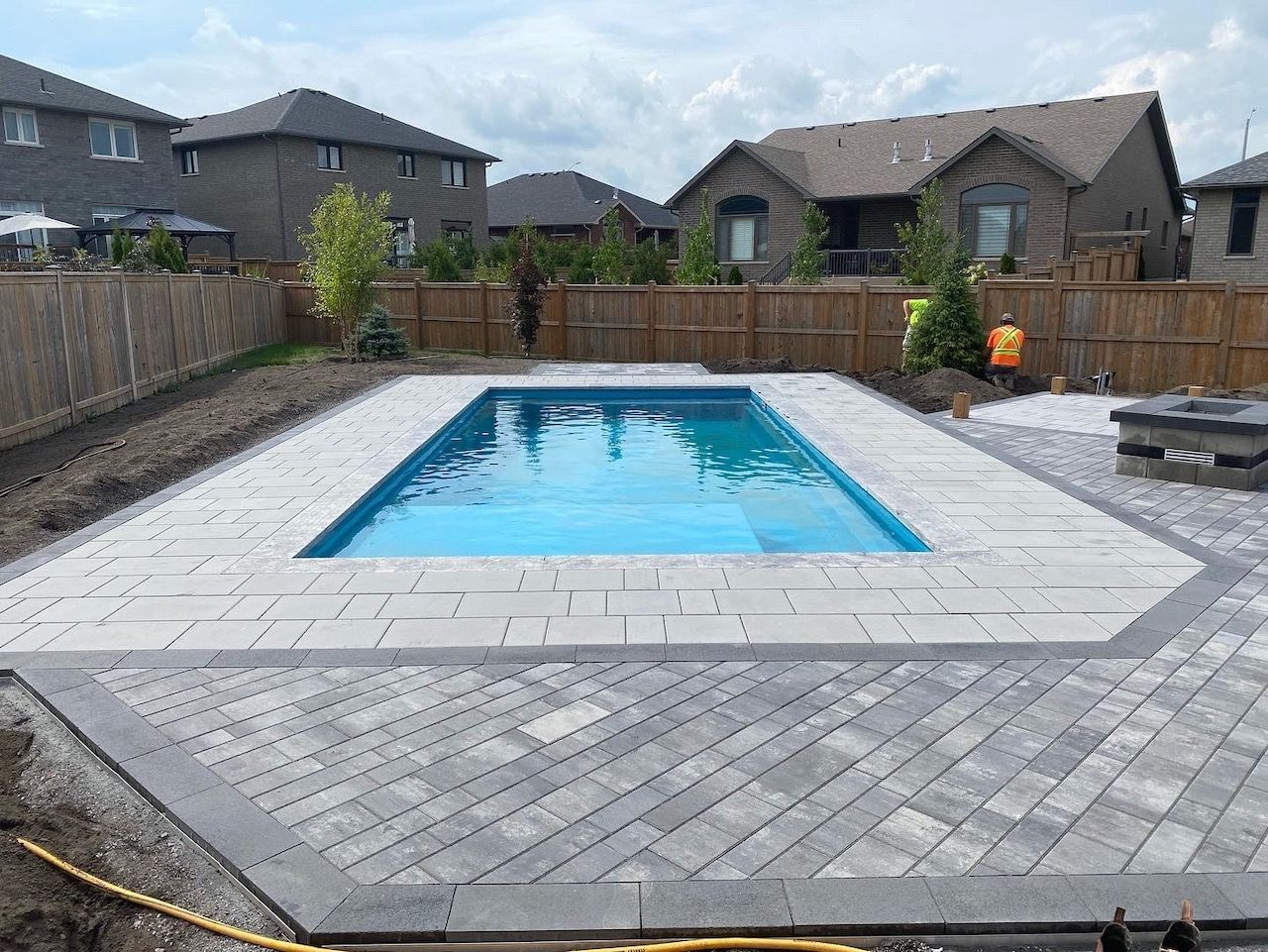 Rectangular in-ground pool surrounded by gray pavers. Backyard setting with houses and a wooden fence.