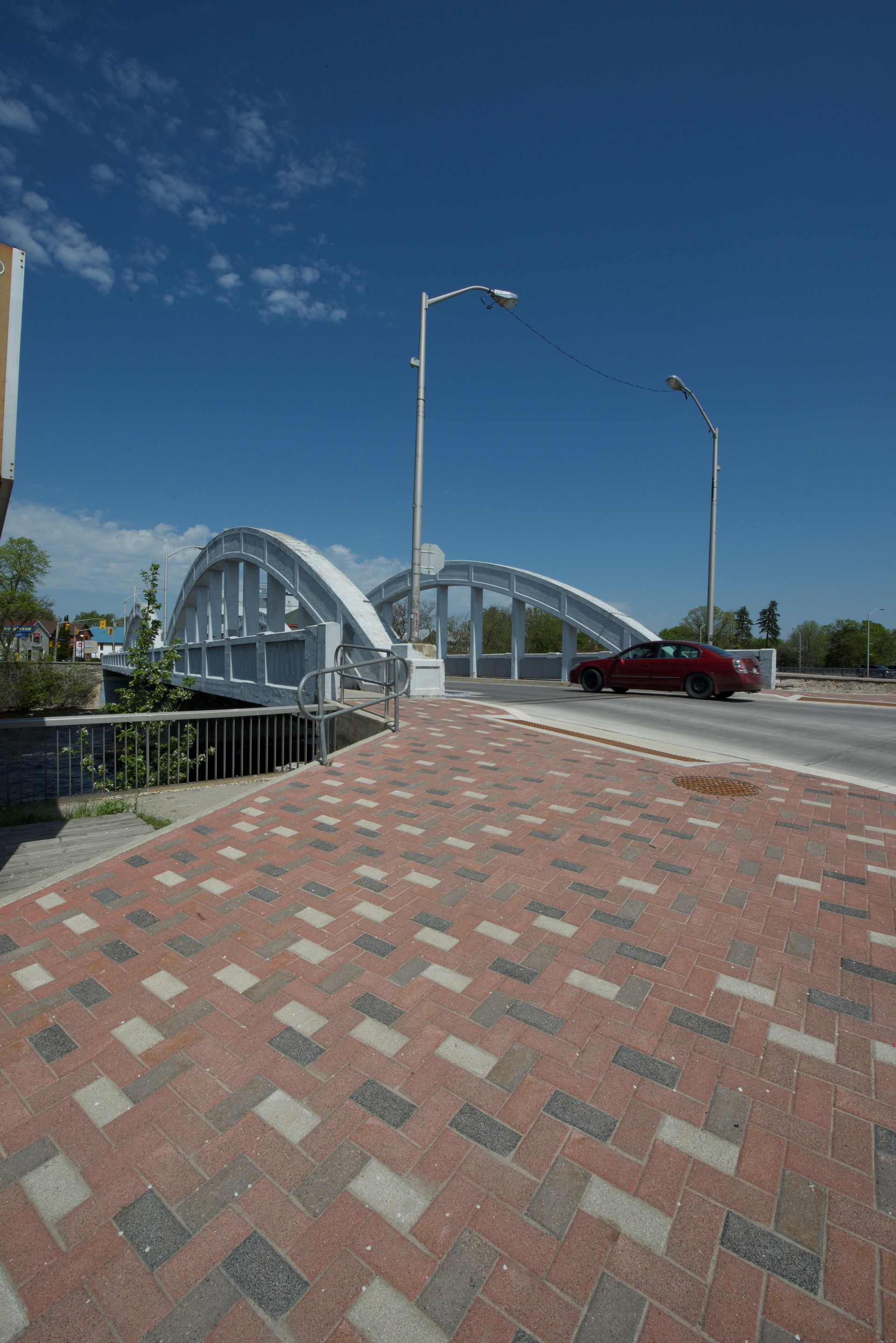 A red car is driving under a bridge on a brick sidewalk.