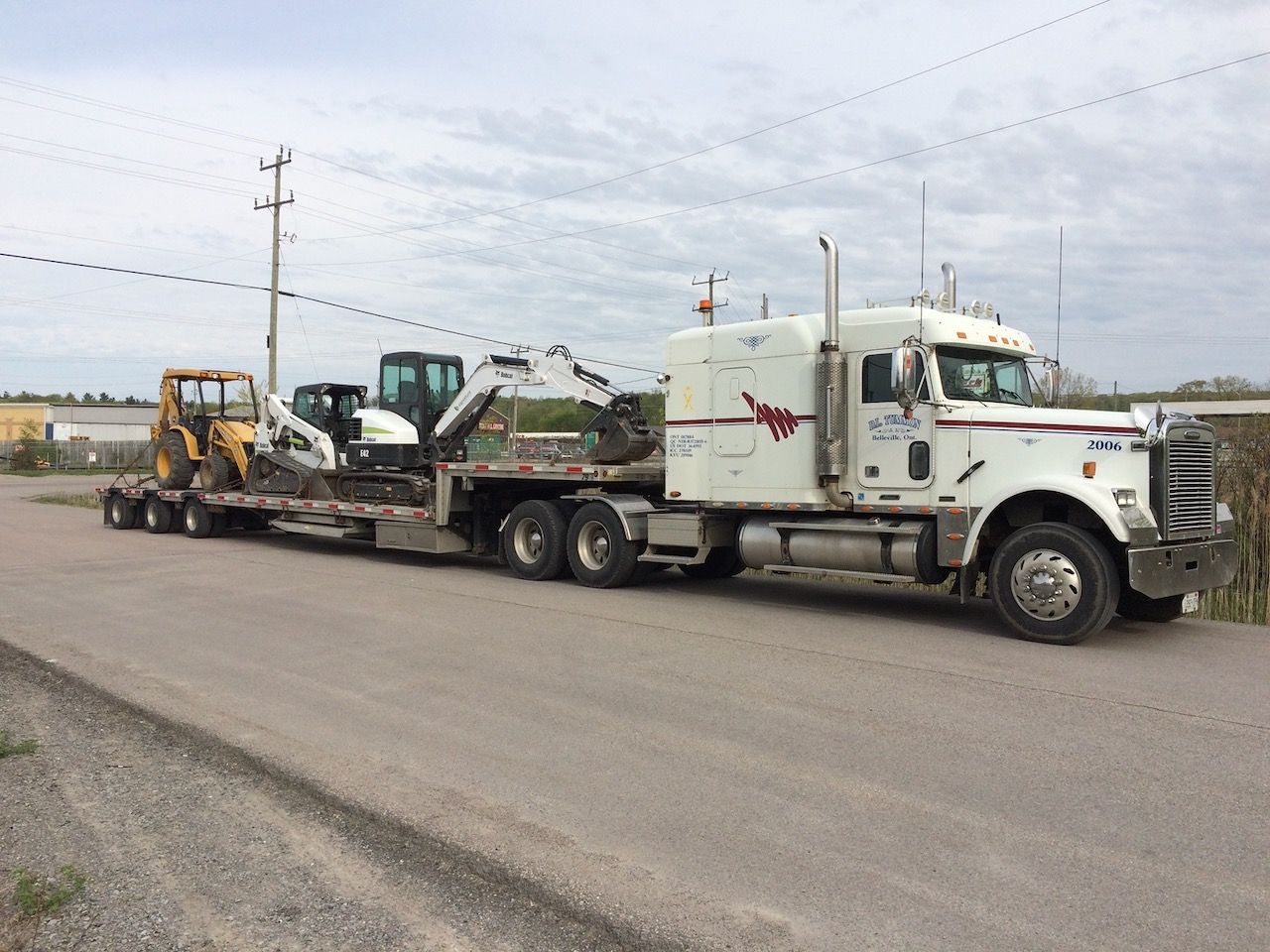 A white semi truck is carrying a yellow excavator on a trailer.