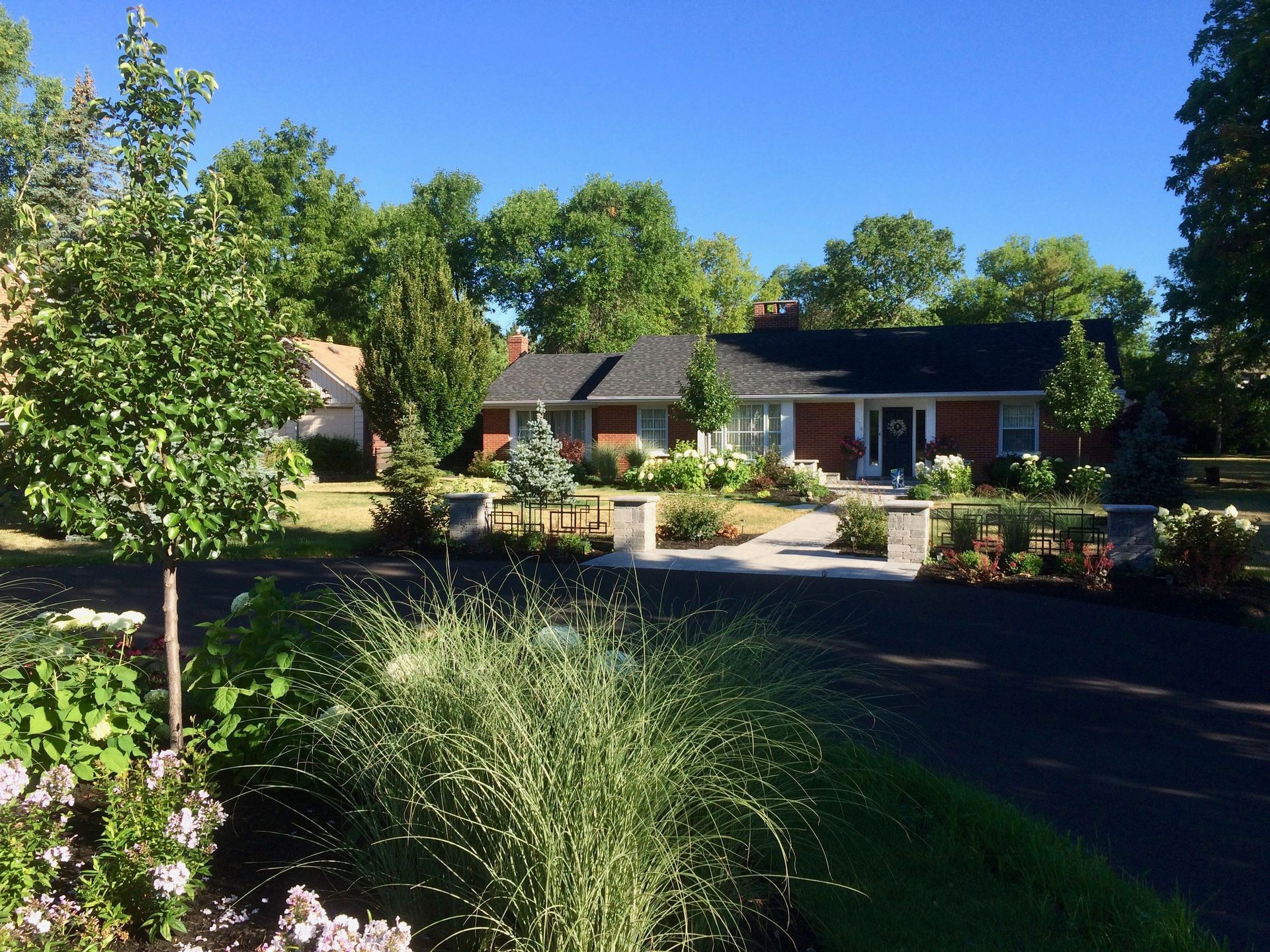 A brick house with a black roof is surrounded by trees and bushes