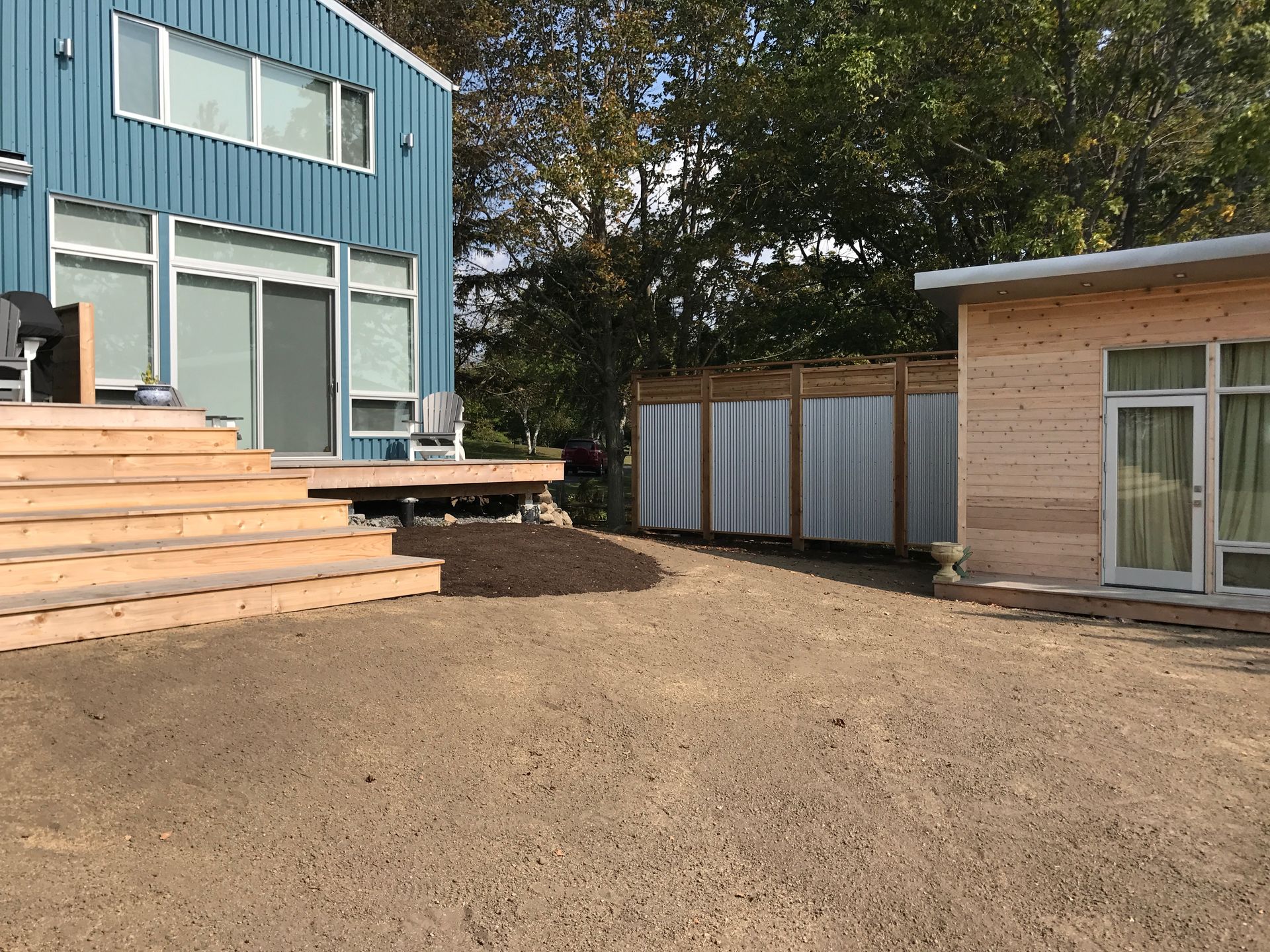 A wooden house with a blue building in the background and a gravel driveway in front of it.