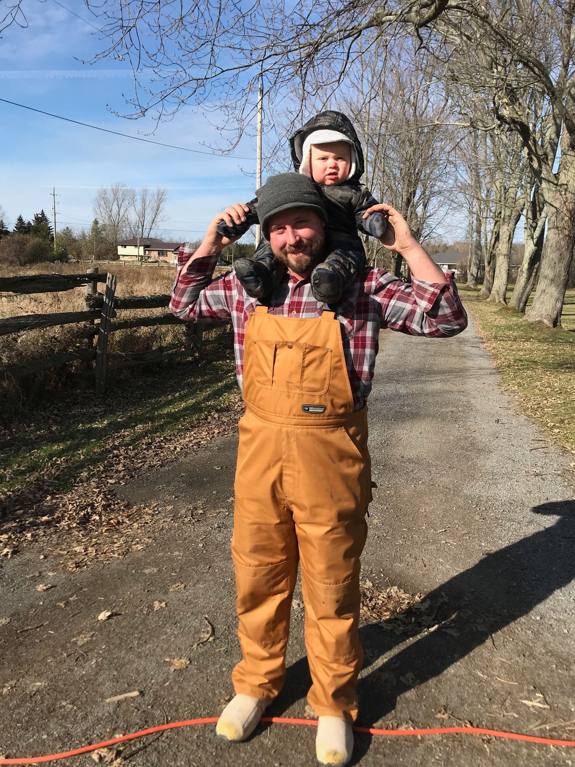 A man in overalls is carrying a baby on his shoulders.