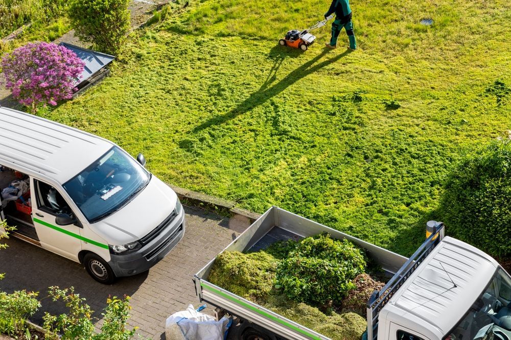 Lawn care service: Person mowing lawn; white van and truck with yard waste; flowers and green grass.