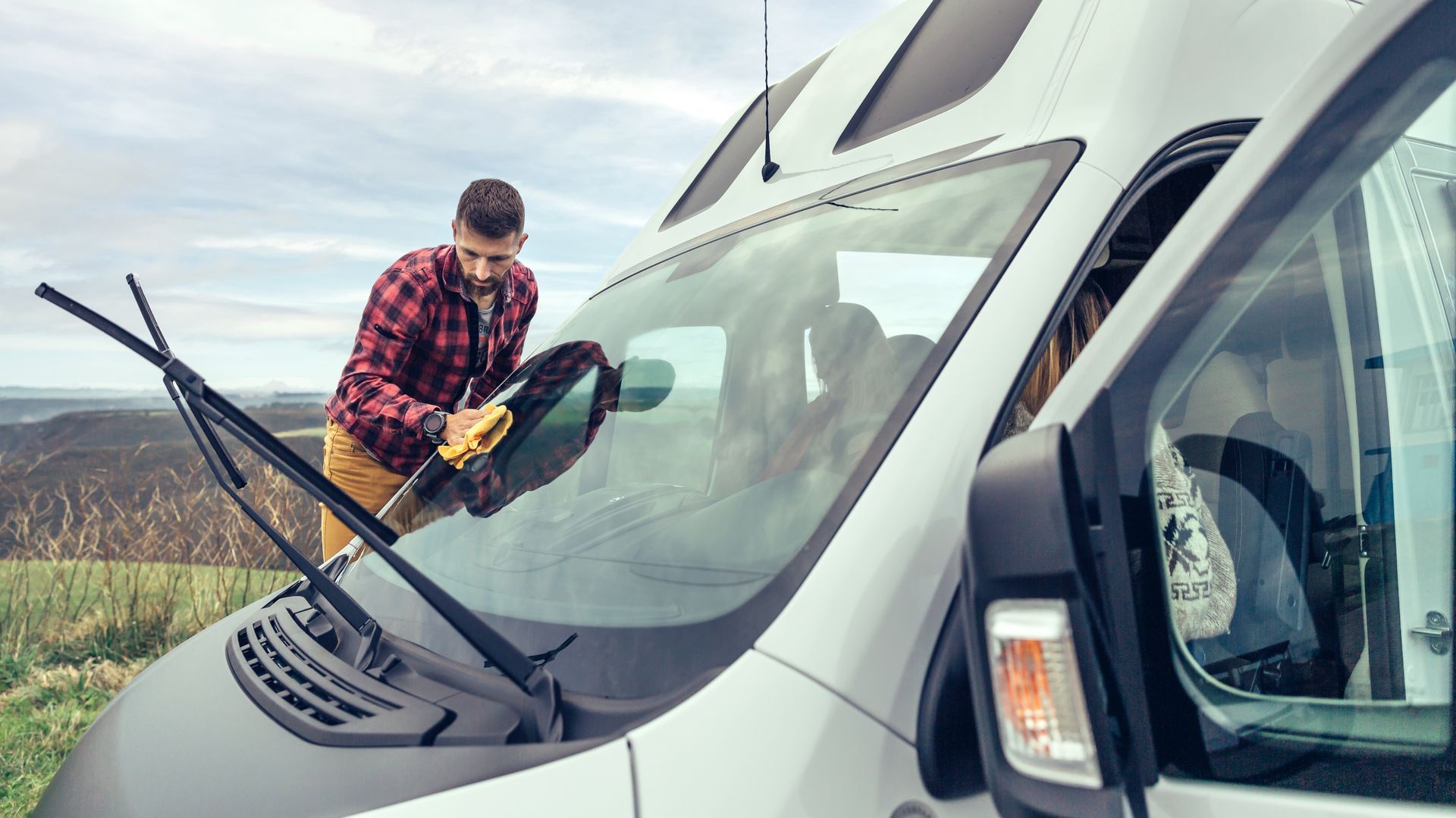 A man uses a piece of cloth to clean the windshield of an RV in the outdoors.