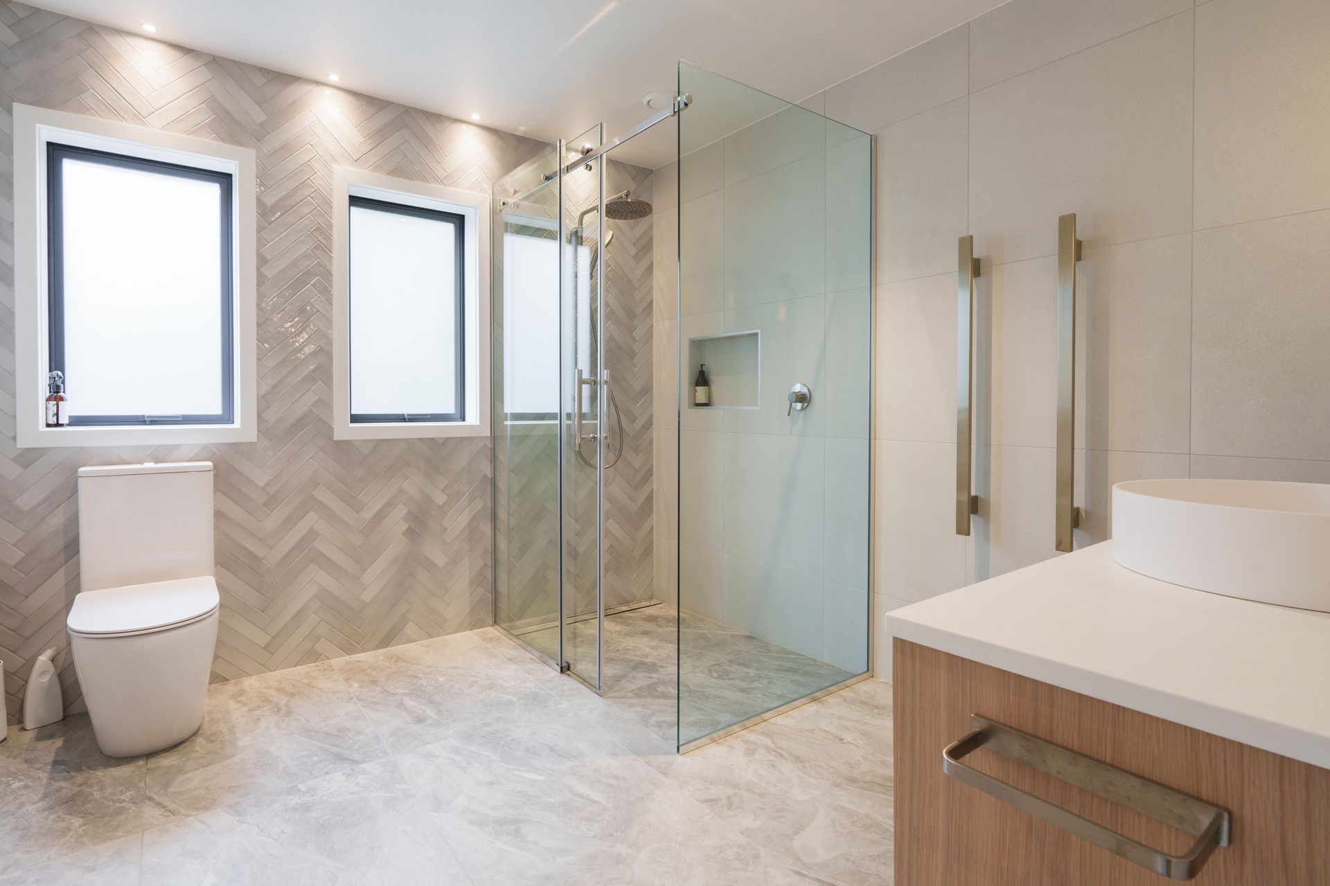 Bright modern bathroom with glass shower enclosure, herringbone wall tiles, and two windows.