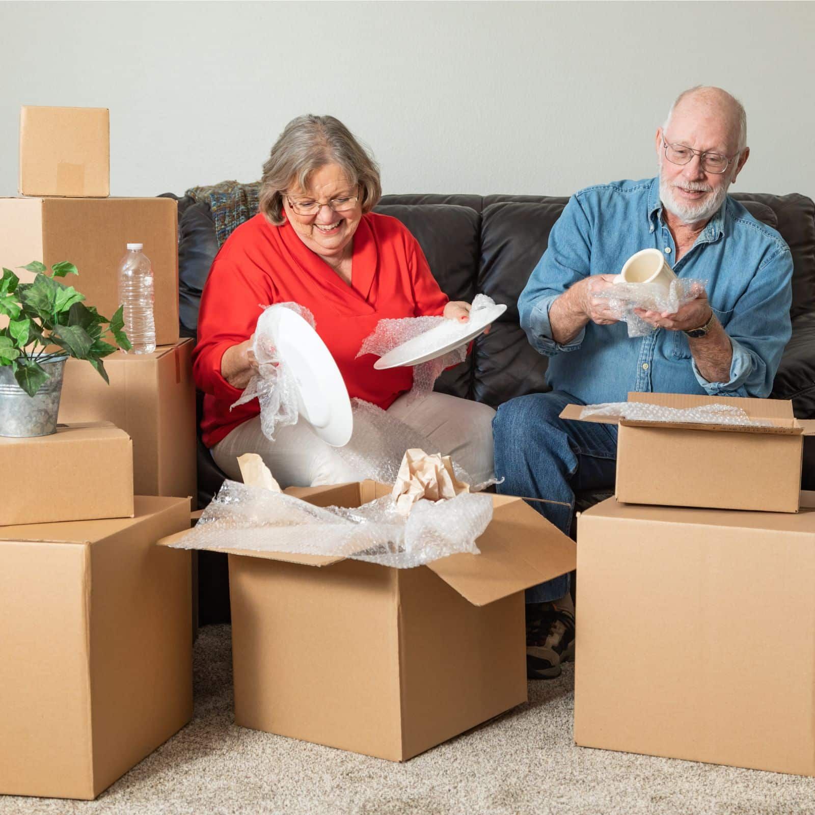 Two people sitting on a sofa and packing plates and a mug into cardboard moving boxes.