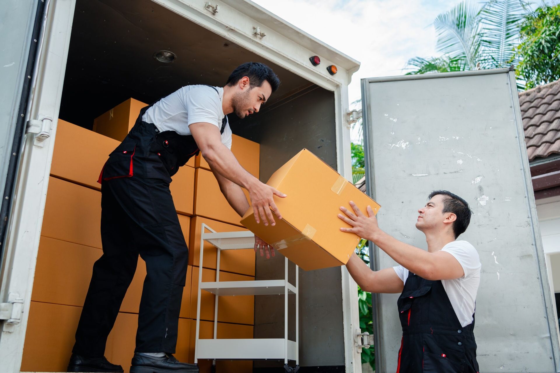 Two movers in uniform unloading cardboard boxes from the back of a delivery truck.