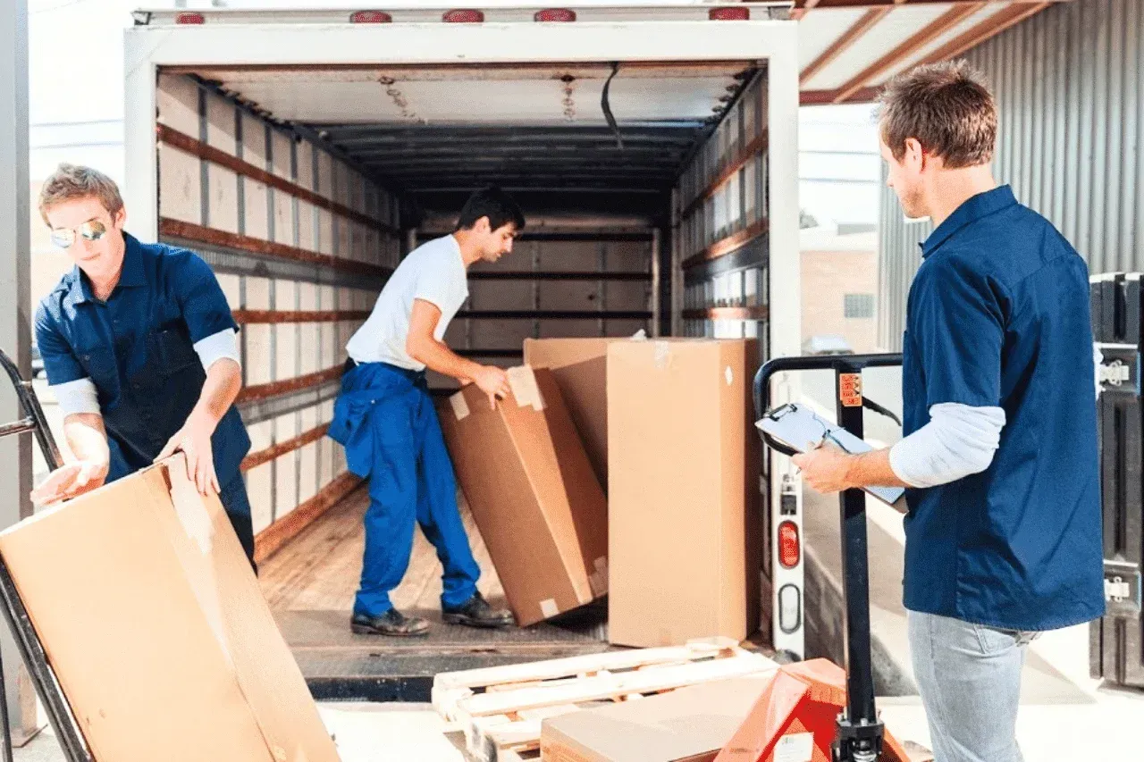 Three workers in blue shirts and pants move cardboard boxes into the back of a moving truck at a loading dock.