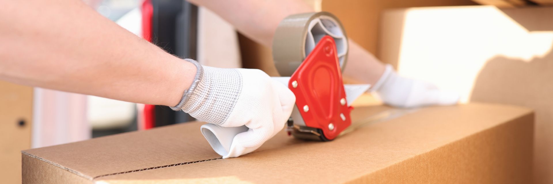 Person wearing white gloves uses a handheld tape dispenser to seal a cardboard box.