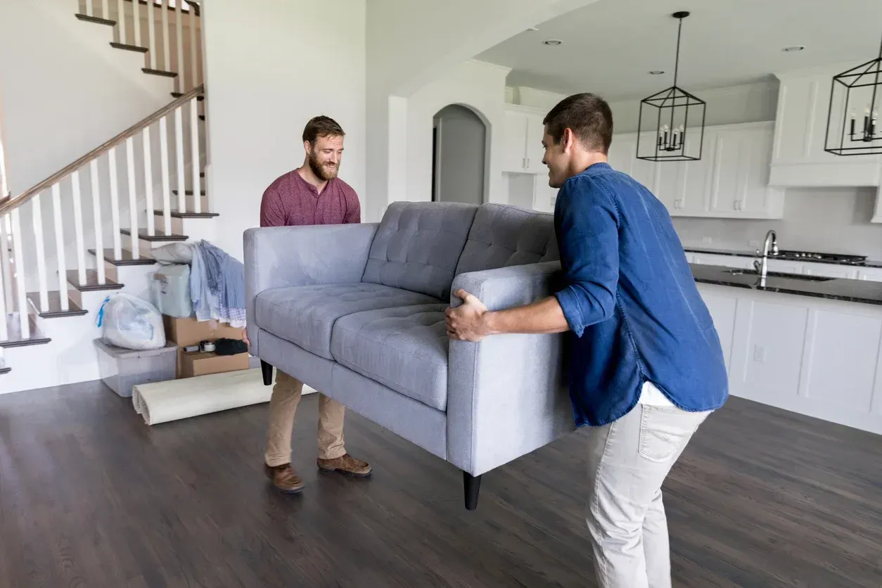 A person and a partner sit on a wood floor, packing belongings into cardboard boxes amidst moving supplies.