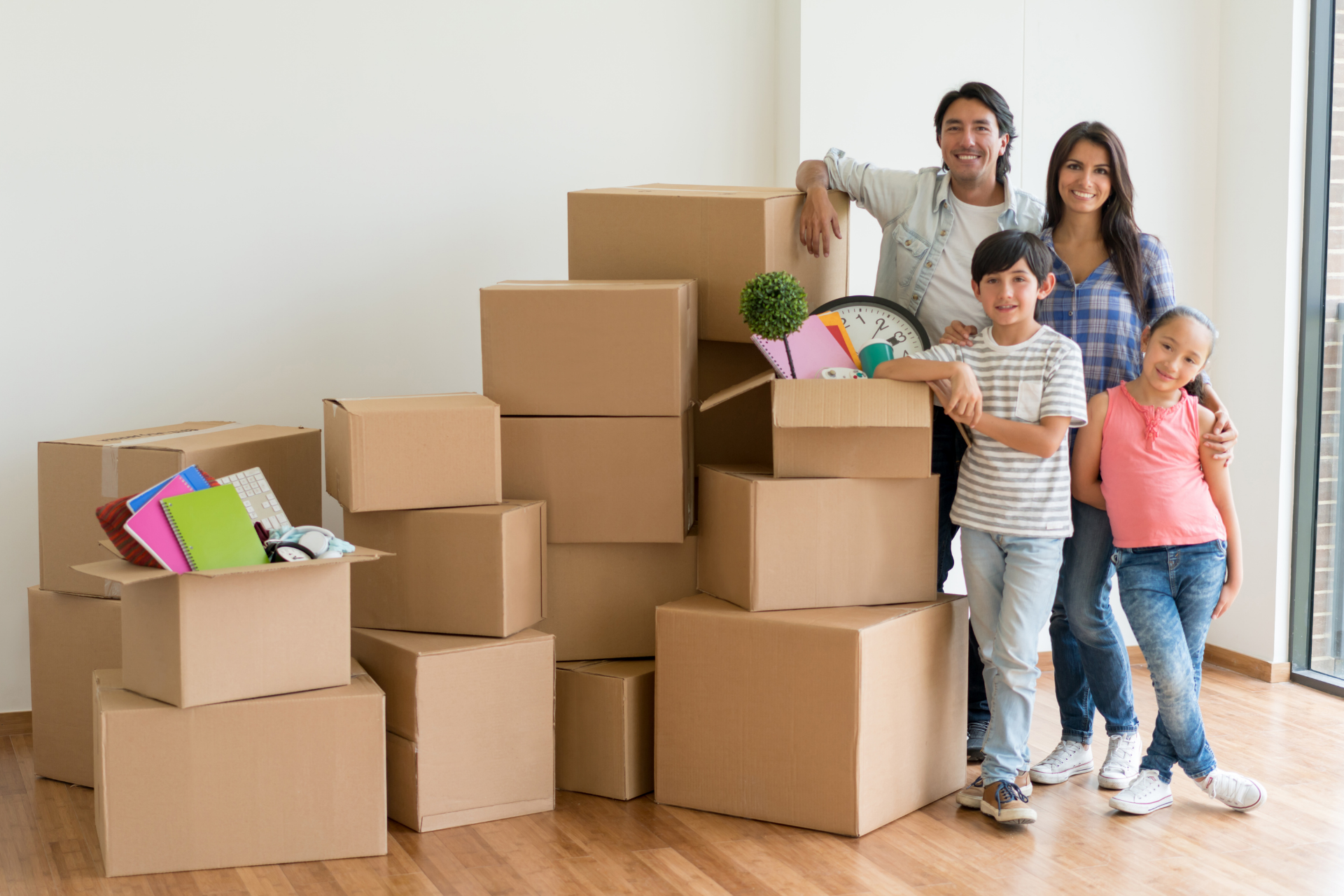 A smiling family stands in an empty room beside a large stack of cardboard moving boxes with colorful notebooks on top.