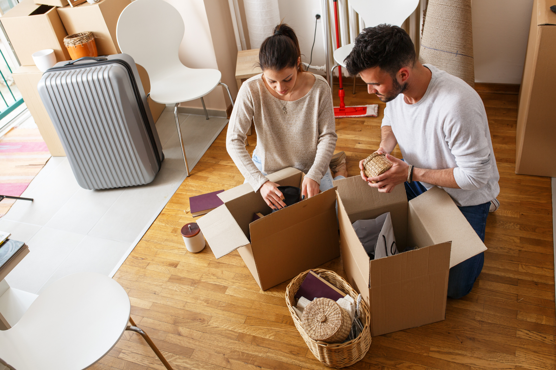 A person and a partner sit on a wood floor, packing belongings into cardboard boxes amidst moving supplies.