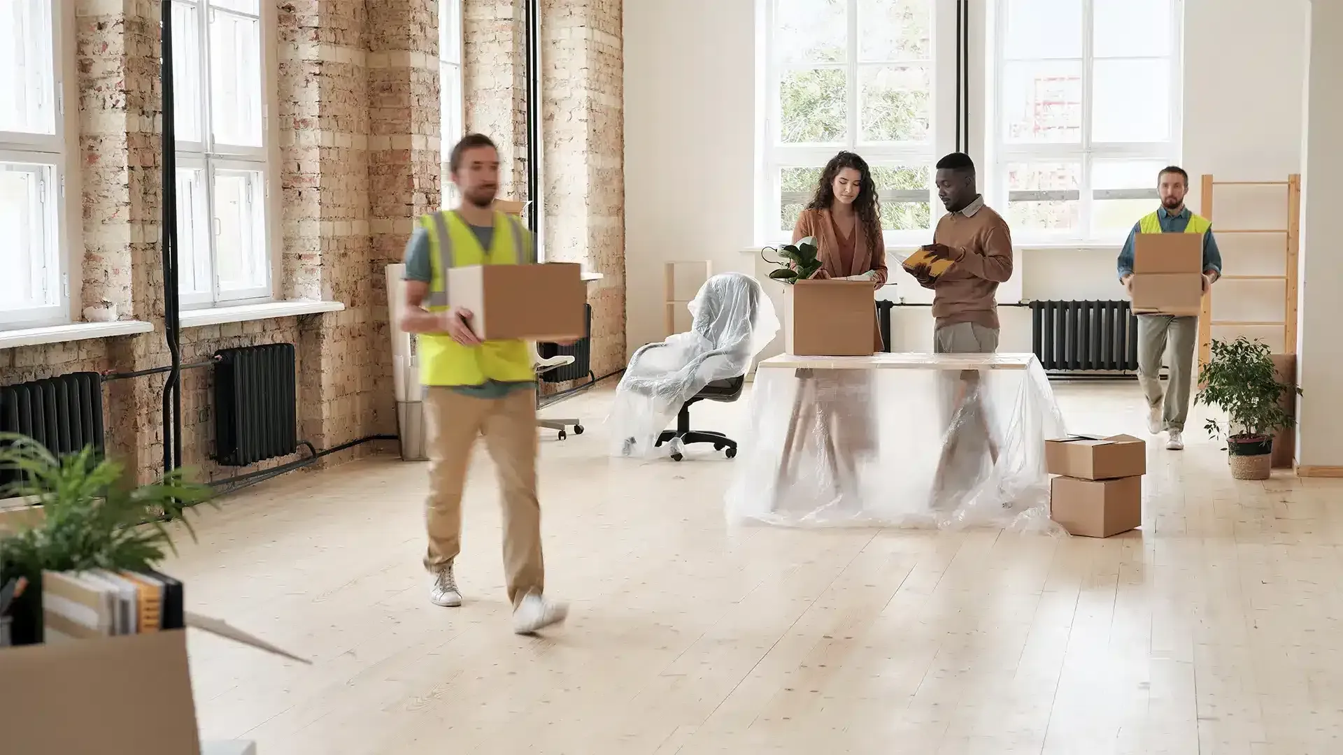 Moving team members in high-visibility vests carry cardboard boxes across a bright, spacious office during relocation.