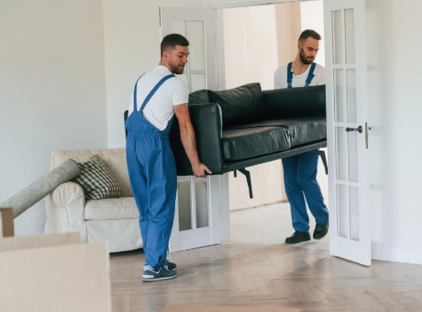 Two movers in blue overalls carrying a black sofa through a doorway into a room with a white armchair and moving boxes.
