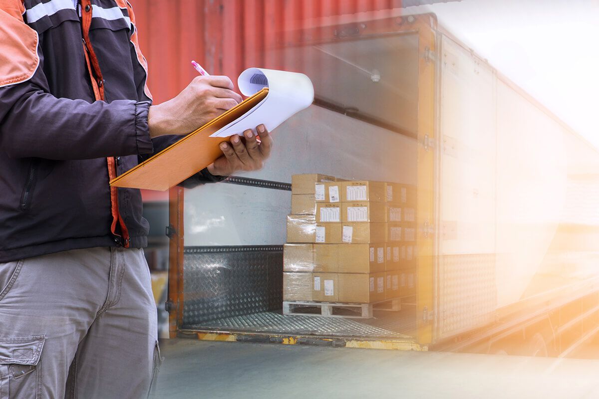 A worker in a jacket holds a clipboard and pen to check shipments inside a warehouse loading bay.