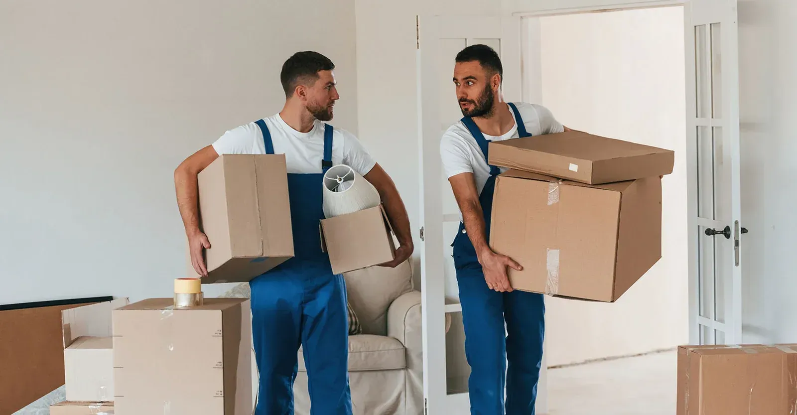 Two movers in blue overalls carrying cardboard boxes and looking at each other in an empty, sunlit room.