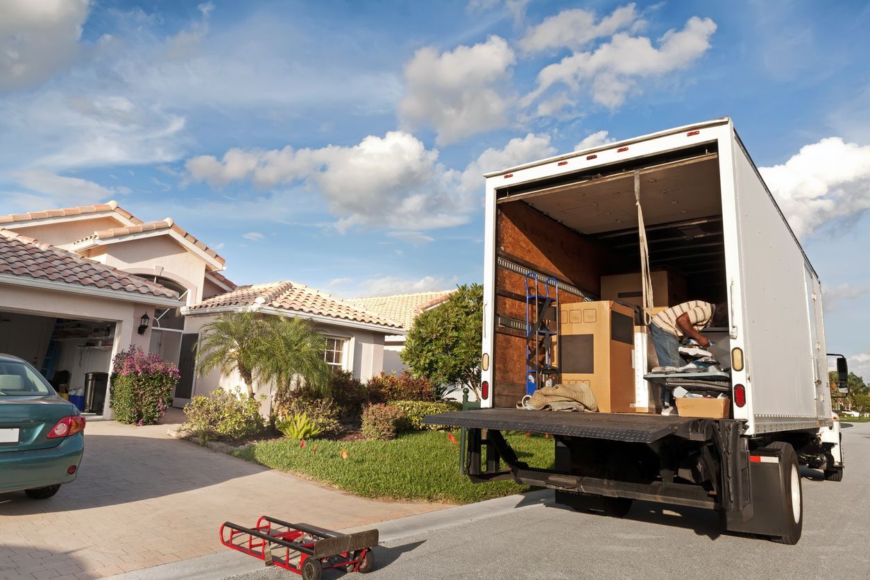 A moving truck with an open cargo door parked on a suburban street in front of a house, with a dolly on the ground.