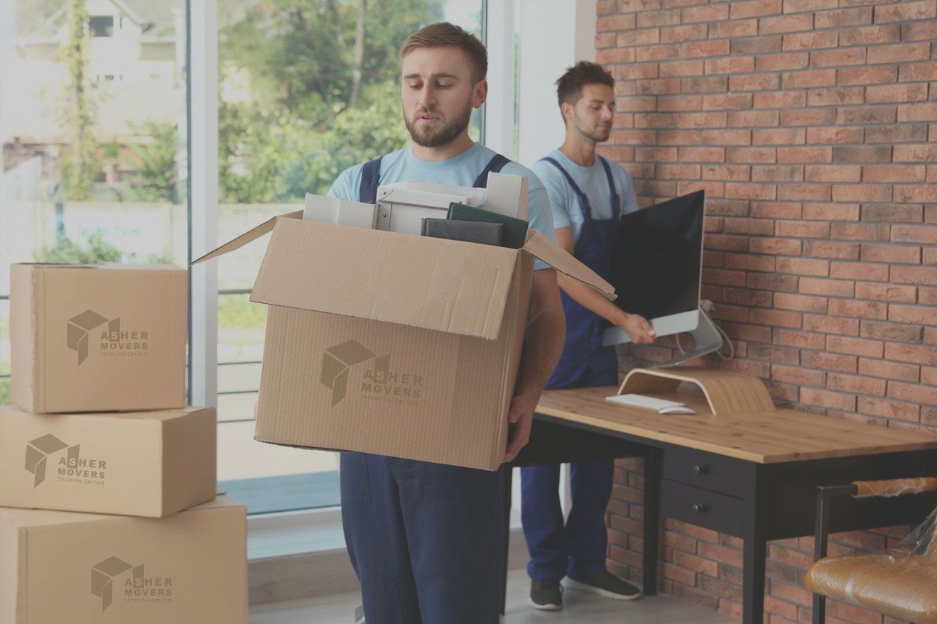Two movers in blue uniforms carry a cardboard box and a computer monitor through an office.