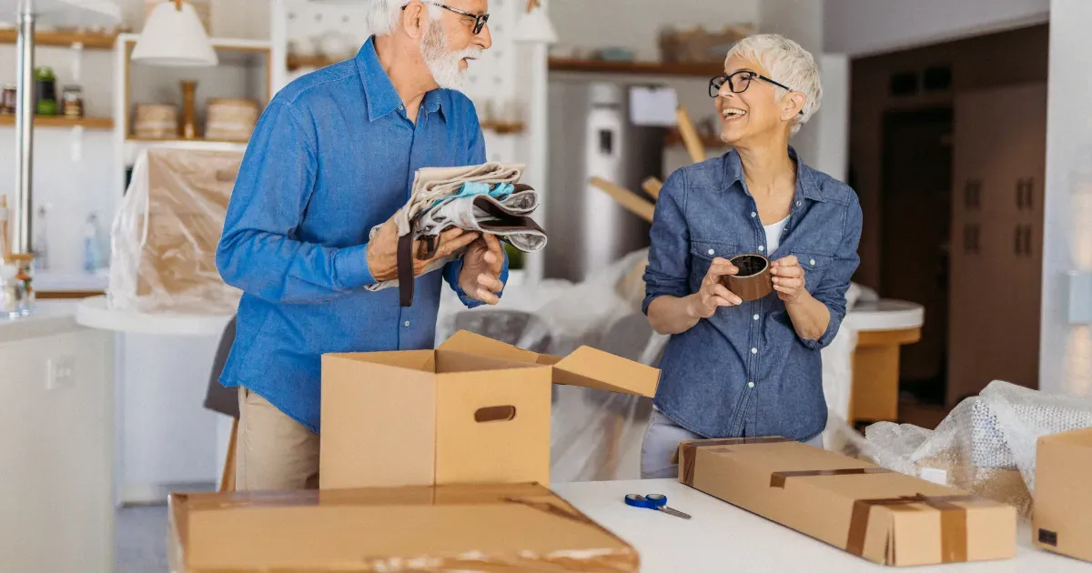 Two people in a kitchen packing items into cardboard boxes while smiling at each other.