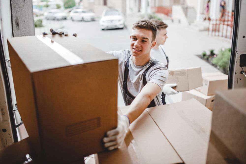 Two delivery workers in uniform loading brown cardboard boxes into the back of a moving truck.
