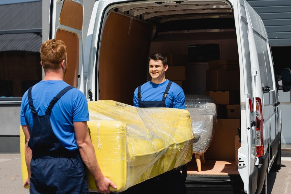 Two movers in blue uniforms and overalls load a yellow, plastic-wrapped sofa into the back of a white van.