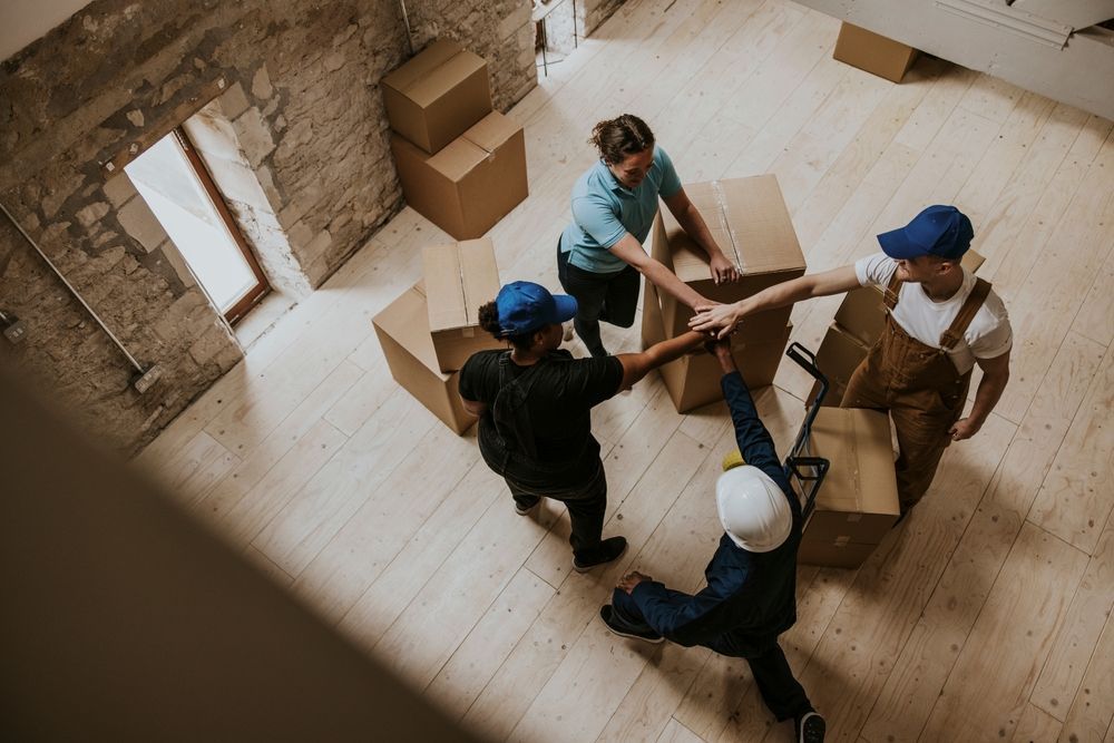 Four workers in a room with cardboard boxes stand in a circle and place their hands together in a sign of teamwork.