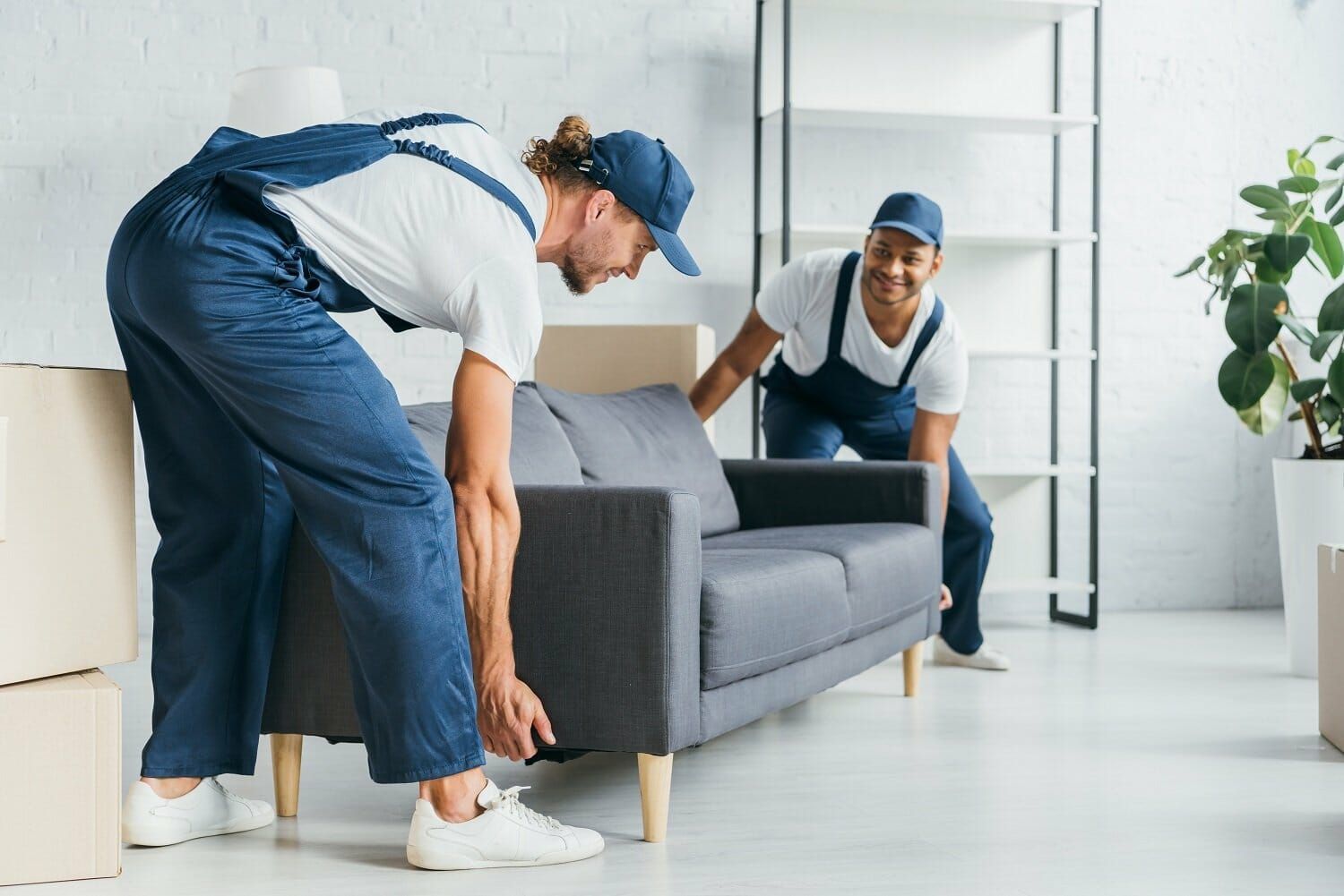 Two movers in white t-shirts and navy overalls carefully lift a grey sofa inside a room with cardboard boxes and shelving.