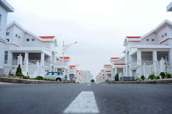 Street lined with matching white houses, red roofs, and a white car. Cloudy sky overhead.