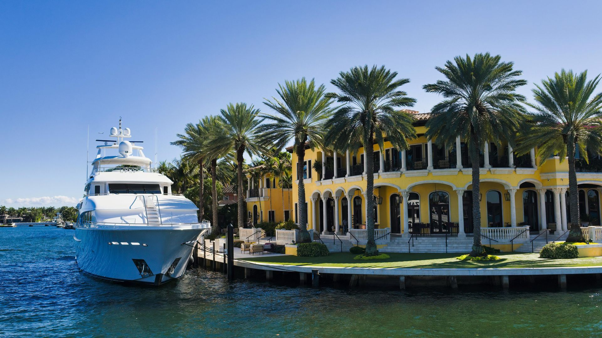 Luxury yacht docked by a yellow mansion with palm trees on a sunny day.