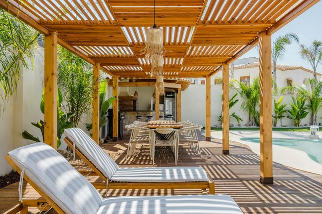 Wooden pergola over lounge chairs and dining table near a pool.