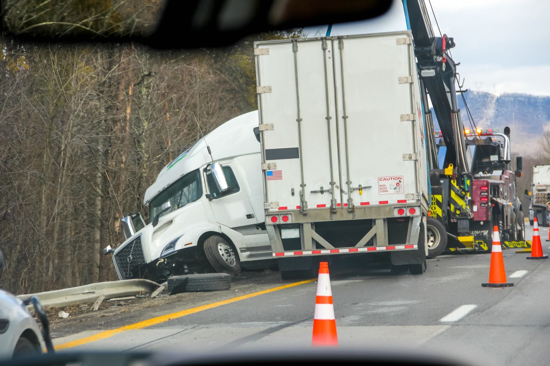 White box truck crashed into a guardrail on a roadside, with orange cones marking the scene.