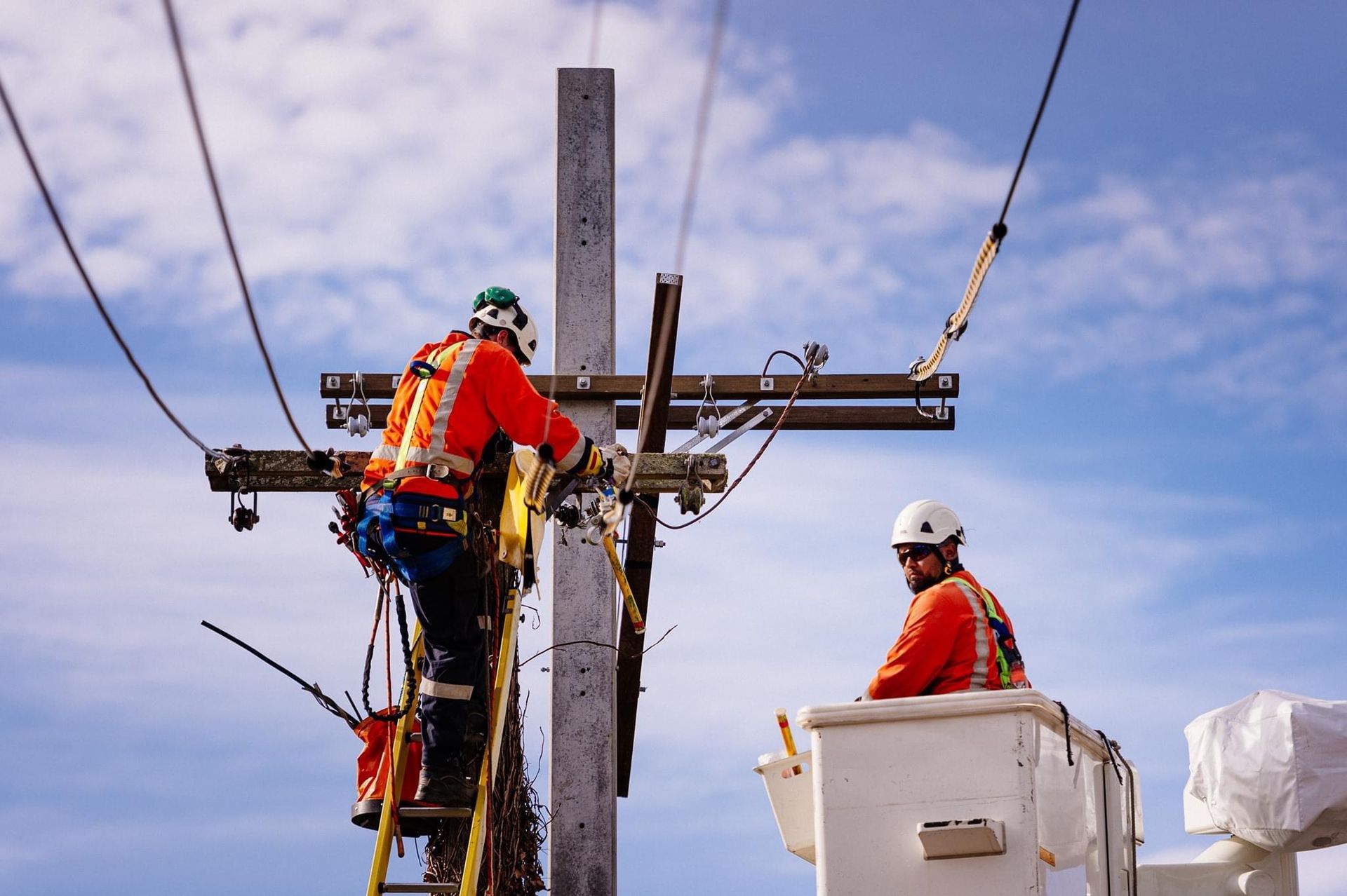 Men in orange vests working on a utility pole, one on a ladder, the other in a bucket lift — L & K Electrical & Air Conditioning in Narrabri, NSW