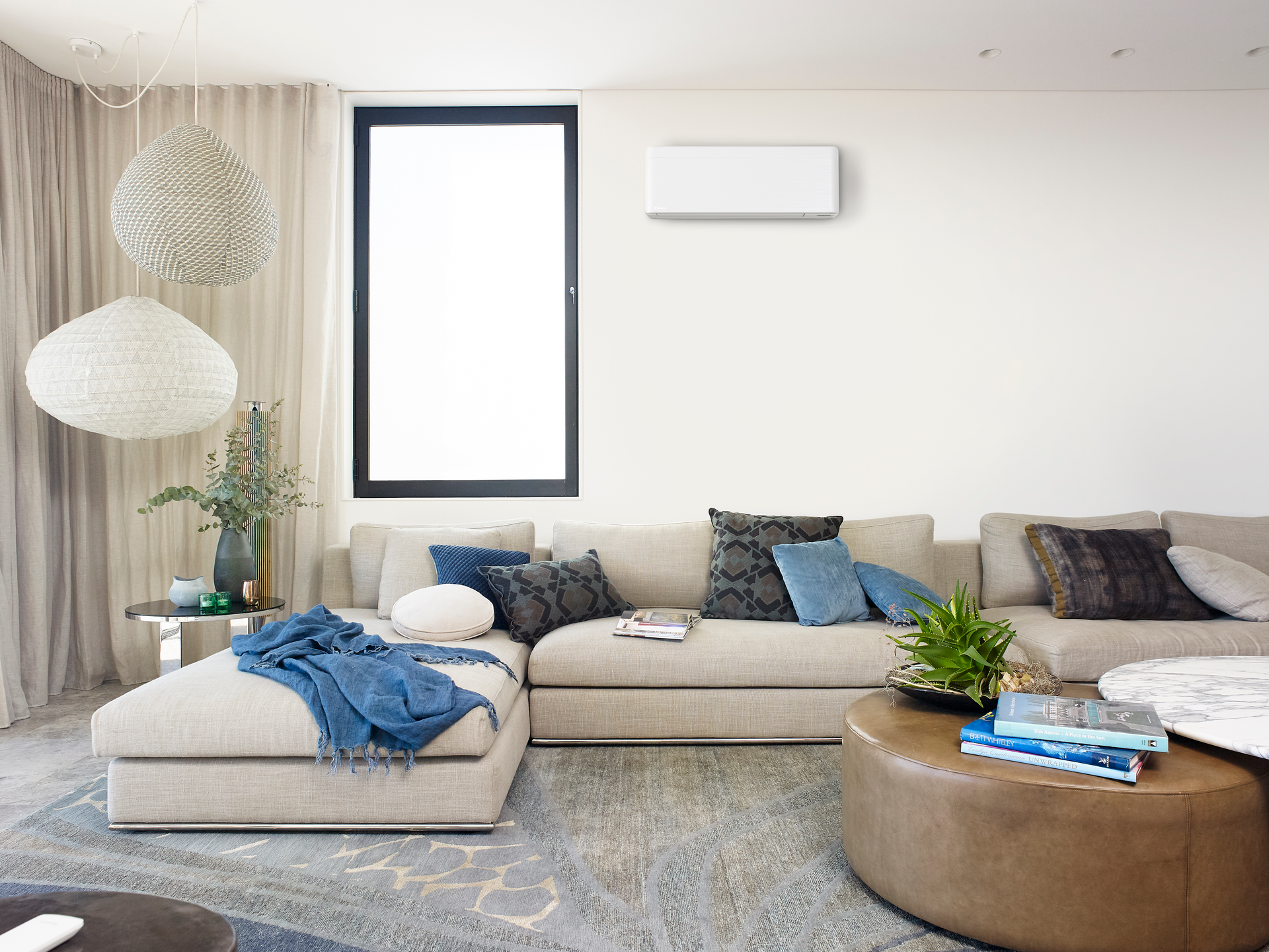 Living room with beige sectional sofa, round ottoman, and air conditioner on white wall — L & K Electrical & Air Conditioning in Uralla, NSW