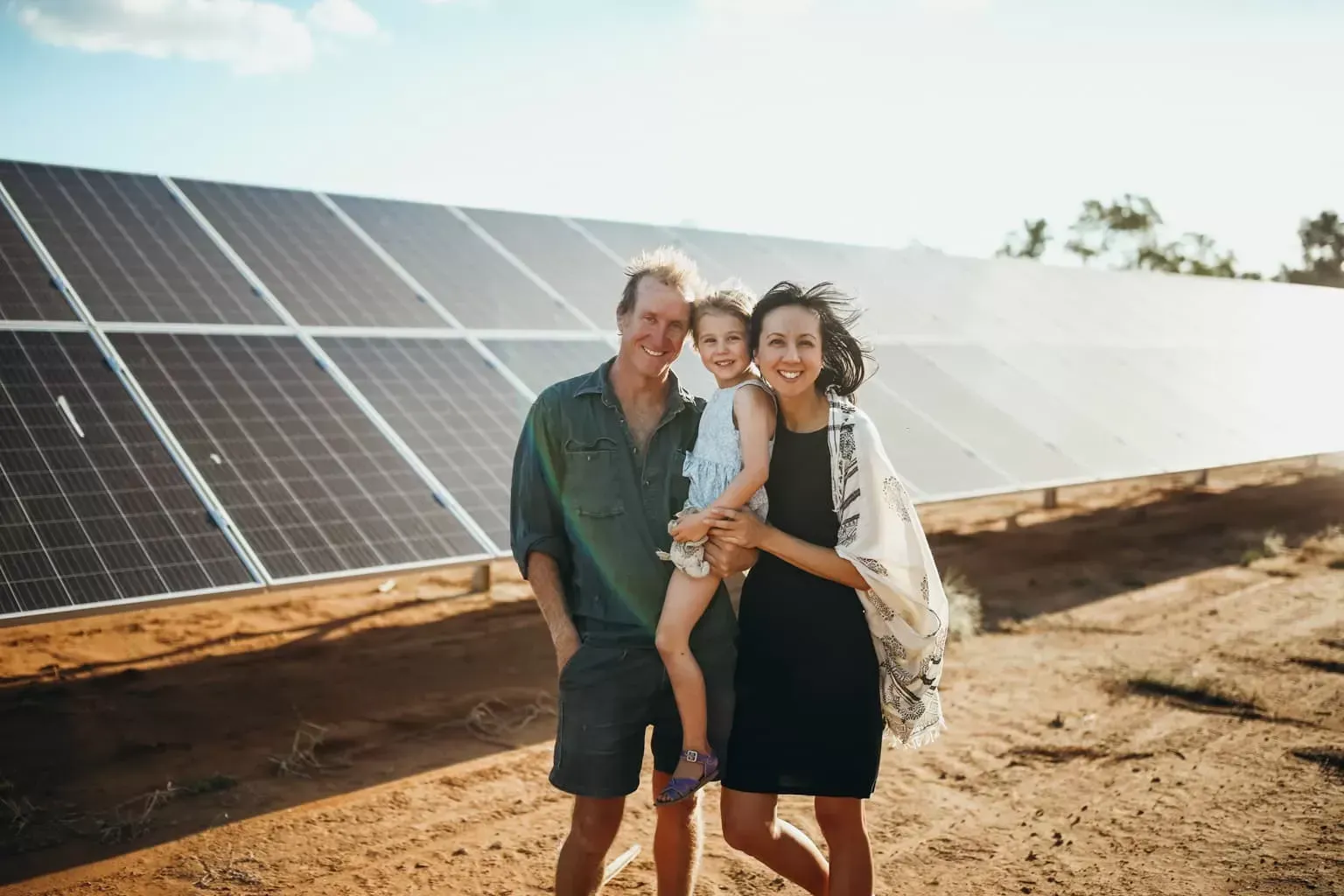 Family of three smiling, posing in front of a solar panel array in a sunny, arid landscape — L & K Electrical & Air Conditioning in Gunnedah, NSW